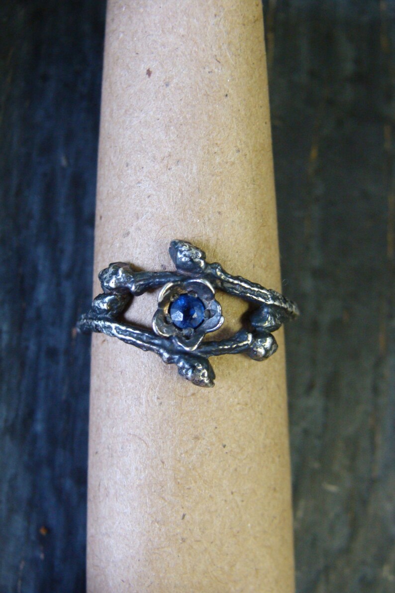 May include: A silver ring with a blue gemstone set in a flower-shaped design. The ring is made to look like branches with small buds.