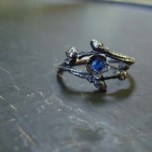 May include: A silver ring with a blue gemstone set in a flower design. The ring is made to look like branches.
