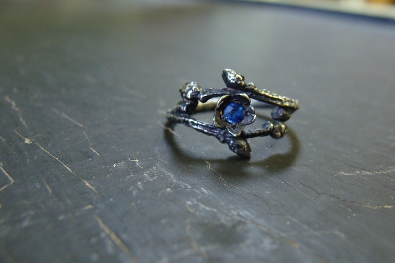 May include: A silver ring with a blue gemstone set in a flower design. The ring is made to look like branches.