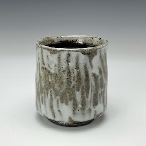 May include: A handmade ceramic cup with a textured, white and gray glaze. The cup has a cylindrical shape with a slightly flared rim and a dark brown interior. The base is a darker shade of brown.