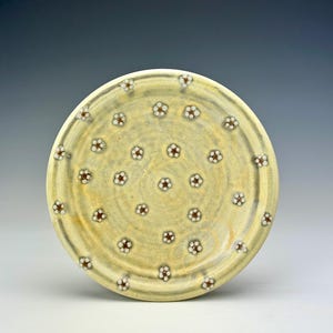 May include: A handmade ceramic plate with a light yellow glaze. The plate is decorated with small, evenly spaced flower designs in white and brown. The plate has a slightly raised rim and measures approximately 10 inches in diameter.