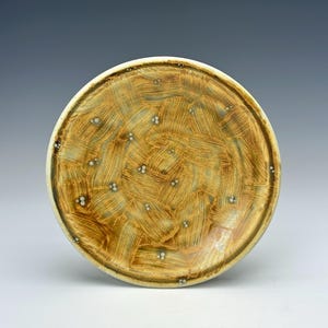 May include: A handmade ceramic plate with a textured surface. The plate features a warm, earthy color palette with brown and tan brushstrokes and small white accents. The rim is a lighter cream color.