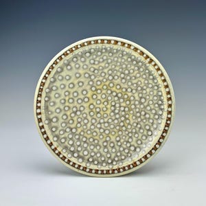 May include: A round ceramic plate with a speckled pattern in shades of white, gray, and yellow. The rim features a border of small brown dots. The plate has a handmade aesthetic.
