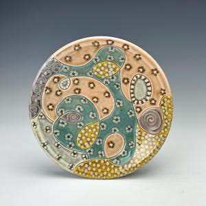 May include: A round ceramic plate with an abstract design. The plate features a light peach background with teal, yellow, and gray organic shapes. The design includes small white flower-like patterns and spiral details.