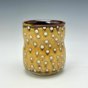 May include: A handmade ceramic tumbler with a textured, golden-brown exterior adorned with white polka dots. The rim is dark brown, and the interior is a deep, glossy brown. This unique, handcrafted cup is perfect for enjoying your favorite beverage.