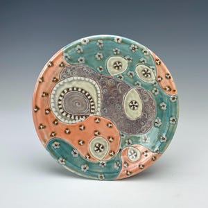 May include: A decorative ceramic plate with a unique design. The plate features a mix of colors, including coral, teal, and purple, with a pattern of abstract shapes and small white flower accents. The plate is round and appears to be handmade.