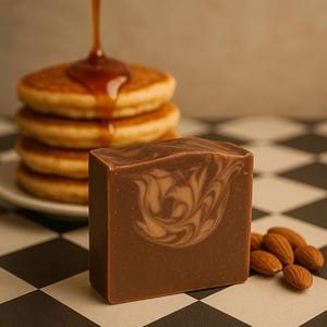 May include: A rectangular bar of brown soap with a light brown swirl pattern, positioned next to a stack of pancakes with syrup being poured over them and a small cluster of whole almonds on a black and white checkered surface.