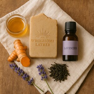 May include: A still-life image featuring natural skincare products. A bar of soap with the text "Wholesome Lather", a small jar of honey, a bottle of lavender essential oil, turmeric root, lavender sprigs, and tea leaves are arranged on a linen cloth.