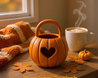 Könnte beinhalten: Ein orangefarbener, kürbisförmiger Korb mit Herz-Ausschnitt und Griff, mit dem Text "Pumpkin Basket", steht auf einer Holzoberfläche. Eine Tasse Kaffee, ein kleiner Kürbis und Herbstblätter bilden eine warme, saisonale Szene.