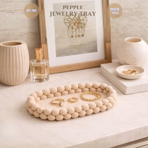 Può includere: Un vassoio ovale beige per gioielli con un bordo di perline, contenente anelli e un braccialetto dorati. Il vassoio è su una superficie bianca, con un'opera d'arte incorniciata e vasi decorativi sullo sfondo. Il testo "PEPPLLE JEWELRY TRAY" è visibile.