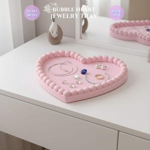 May include: A pink heart-shaped jewelry tray with a beaded border. The tray holds various jewelry items, including a blue gemstone, a gold ring, and several clear crystals. The text "BUBBLE HEART JEWELRY TRAY" is visible.