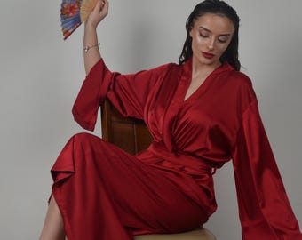 May include: A woman wearing a long, red satin robe with a V-neck and a sash. She is sitting on a wooden chair and holding a blue and gold hand fan.