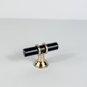May include: A cabinet pull with a black cylindrical handle and a gold-colored base. The handle is held in place by two gold-colored rings. The base has a rounded shape. This hardware is suitable for drawers and cabinets.