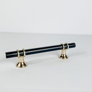 May include: A black cabinet pull with gold-colored accents. The handle is a straight bar, and the base is a decorative, rounded shape. The hardware is designed for use on drawers or cabinets.