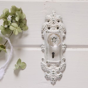 May include: Ornate white door handle with a clear glass knob. The handle has a distressed finish, with hints of green peeking through the white paint. Dried green flowers and white ribbon are in the background.