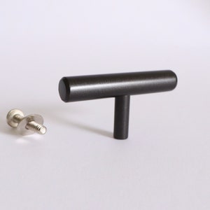 May include: A dark gray T-bar cabinet pull with a matching screw and washer. The hardware is made of metal and is designed for use on drawers and cabinets. The T-bar handle is cylindrical.