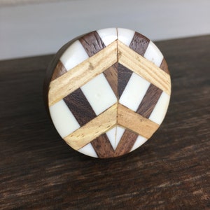 May include: A round wooden cabinet knob with a geometric design. The knob features alternating sections of dark brown wood, light wood, and white material. The knob is set against a dark brown wooden surface.