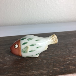May include: A ceramic fish figurine with a white body, green spots, and a reddish-brown head. The tail is tan. The fish has a single, large eye and is resting on a wooden surface. The figurine is a decorative item.