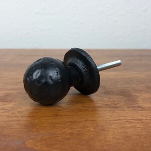 Puede incluir: Pomo de armario de metal negro con un dise&ntilde;o redondo y texturado. El pomo tiene una base plana y un tornillo para fijarlo a una puerta de armario.