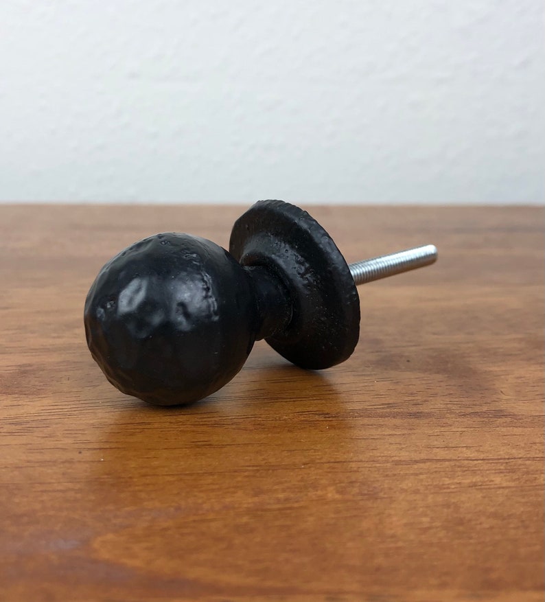Puede incluir: Pomo de armario de metal negro con un dise&ntilde;o redondo y texturado. El pomo tiene una base plana y un tornillo para fijarlo a una puerta de armario.