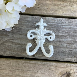 May include: A distressed white metal wall hook with a fleur-de-lis design. The hook has two curved arms and two holes for mounting. The hook is set against a weathered wood background, with white flowers in the upper left corner.