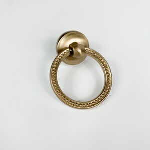 May include: A bronze-colored cabinet pull with a circular handle and a decorative beaded edge. The pull has a round backplate and a ring-shaped handle. The hardware is designed for use on drawers or cabinet doors.