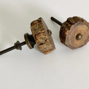 May include: Two rustic wooden cabinet knobs. One is square, the other is round. Both have a natural bark edge and a brass-colored metal base and screw. The wood grain is visible on the round knob.