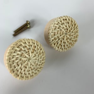 May include: Two round cabinet knobs with a woven rattan design. The knobs are light beige and have a natural wood base. Two gold-colored screws are also visible.