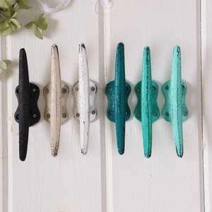 May include: Six decorative cleat hooks in a row, mounted on a white wooden surface. The hooks are painted in various colors: black, cream, white, teal, turquoise, and light turquoise. The hooks are made of metal.