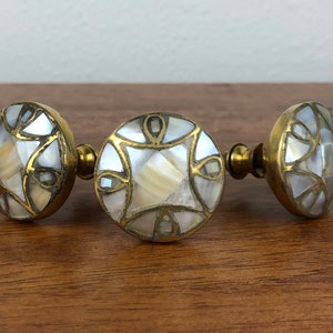 May include: Three decorative cabinet knobs with a round, brass base and a mosaic design of white and cream-colored shell pieces. The knobs are arranged horizontally on a wooden surface.