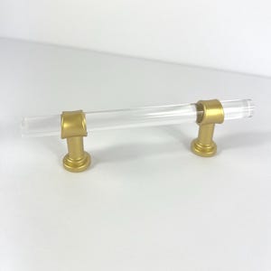 May include: A clear acrylic cabinet pull with gold-colored metal accents. The pull has a cylindrical handle and is supported by two gold-colored posts. The hardware is designed for use on drawers and cabinets.
