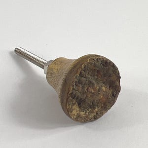 May include: A wooden cabinet knob with a textured, bark-like surface. The knob is a light brown color with darker brown and tan accents. A silver-colored threaded rod extends from the back of the knob. The knob is designed for use on cabinets and drawers.