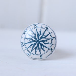 May include: White ceramic knob with a blue compass design. The compass features a star with points extending to the cardinal directions: N, NE, E, SE, S, SW, W, NW.