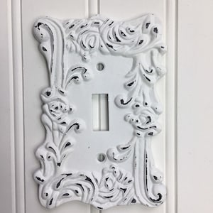 May include: White, ornate light switch cover with a distressed finish. The cover features a rectangular shape with a toggle switch opening and two screw holes. The decorative frame has floral and scroll details.