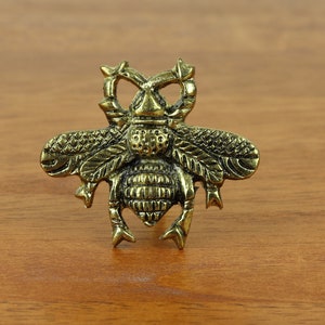 May include: A gold-toned metal bee figurine with intricate details. The bee has a textured body and wings, and its legs are curled up.