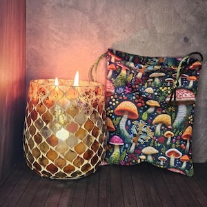 May include: A lit candle in a mosaic-style glass jar next to a drawstring bag. The bag features a colourful mushroom pattern on a dark blue background. The candle's flame glows warmly, illuminating the jar's details.
