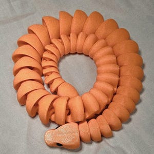 Puede incluir: Un juguete naranja con forma de serpiente segmentada, enrollado en un patrón circular. El juguete tiene una superficie texturizada y una cabeza detallada con un pequeño ojo negro. Los segmentos están curvados, dando al juguete una apariencia flexible.