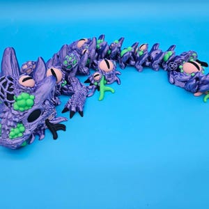 May include: A purple articulated dragon figure with a textured, scaly surface, featuring bright green circular accents, black clawed feet, and multiple pinkish-tan eyes embedded along its long, segmented, winding body on a blue background.