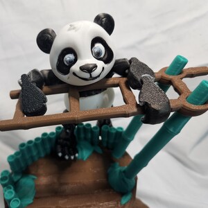 Peut inclure: Figurine de panda imprimée en 3D, avec une échelle et des accents de bambou. Le panda est noir et blanc avec des yeux bleus et tient une échelle marron. Des tiges et des feuilles de bambou vertes sont également présentes.