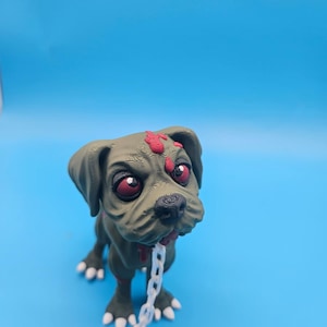 以下が含まれることがあります： 3Dプリントされたオリーブグリーンの犬のフィギュア。表面は質感があり、大きな赤い目、額の赤い斑点、首輪に取り付けられた白いプラスチック製のチェーンが特徴です。犬はシワのある顔と尖った白い爪をしており、深刻な表情を浮かべています。