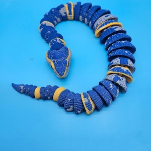 May include: A segmented, articulated toy snake featuring a textured surface with a color palette of deep blue, bright yellow, and light gray. The snake is coiled in a curved position, displaying a detailed head and a tapering tail section.