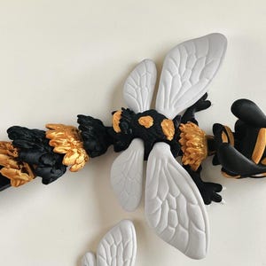 May include: A handcrafted, decorative bee figurine. The bee features a black body with gold and white accents. The wings are white with detailed vein patterns. The bee's abdomen and head have gold and black details.