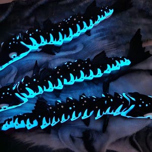 May include: Three articulated fish figures in black with glowing cyan bioluminescent-style markings along their dorsal ridges, heads, and fins. Each fish features a segmented body design with distinct light-emitting patterns on the dark surface.