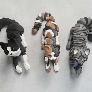 May include: Three handmade cat figurines. One is black and white, another is calico, and the third is grey with stripes. Each cat has a curved tail and is lying down.