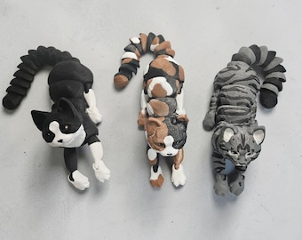 May include: Three handmade cat figurines in various colors. One is black and white, another is calico, and the third is gray with stripes. Each cat has a curved tail and is lying down.
