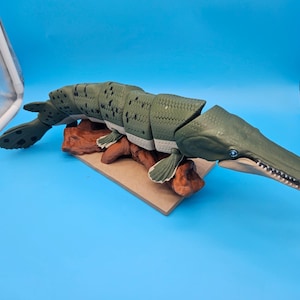 Puede incluir: Una figura articulada de pez catán verde con cuerpo texturizado y segmentado, manchas oscuras y un hocico largo con dientes visibles. El pez tiene el vientre claro y ojos azules, y está montado sobre una base marrón que imita madera.