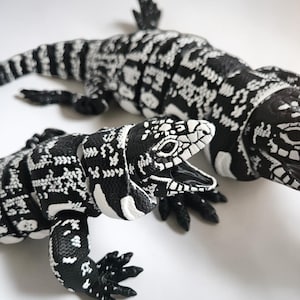 May include: Two articulated lizard figurines featuring a high-contrast black and white pattern across their bodies. The smaller lizard in the foreground has an open mouth, while the larger lizard is positioned behind it, both showing detailed scales.