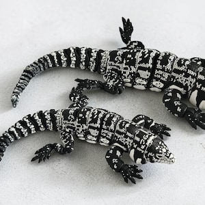 May include: Two detailed lizard figurines in black and white, with a scale-like pattern. The reptiles are positioned on a light grey surface, showcasing their intricate design and realistic features. The lizards have small, detailed feet and long tails.