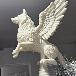 Puede incluir: Figura blanca y detallada de un lobo alado, posiblemente un adorno para el árbol de Navidad. El lobo tiene grandes alas emplumadas y patrones intrincados en su cuerpo. La figura está montada sobre una base verde y plateada.