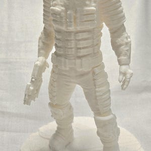 May include: A white 3D-printed figurine of a space suit-clad character, standing on a circular base. The figure has a detailed, textured surface, with visible armor plating and a helmet. The figurine is approximately 7 inches tall.